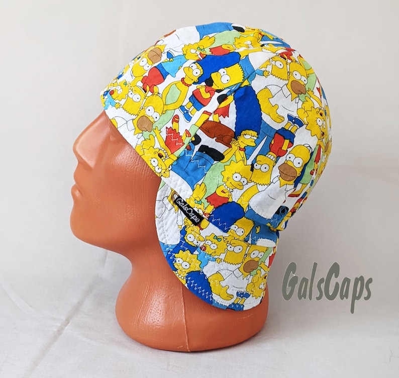 May include: A white welding cap with a blue and yellow pattern featuring characters from the Simpsons cartoon. The cap has a flap that can be worn down to protect the neck and ears. The brand name "GalsCaps" is printed on the flap.