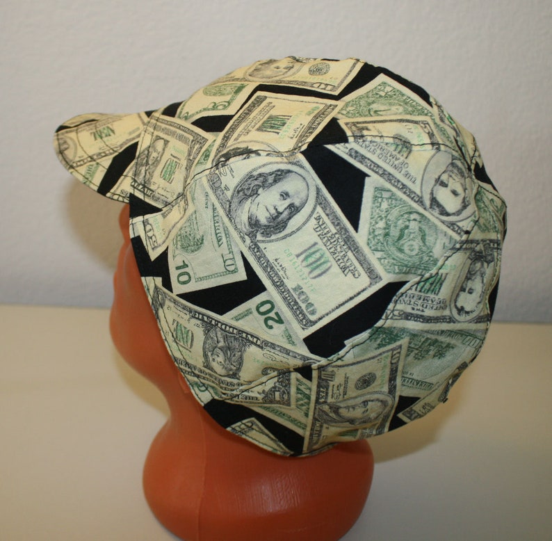 May include: A cap featuring a repeating pattern of US dollar bills in shades of green, black, and cream. The cap has a curved brim and is made of fabric. The design includes images of George Washington and other details from the currency.