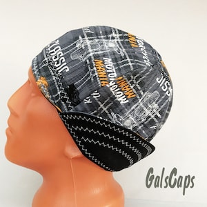 May include: A black and gray patterned beanie with ear flaps. The beanie has the text "MANI MANIA MOTORCYCLE CLASSIC" printed on it in orange.