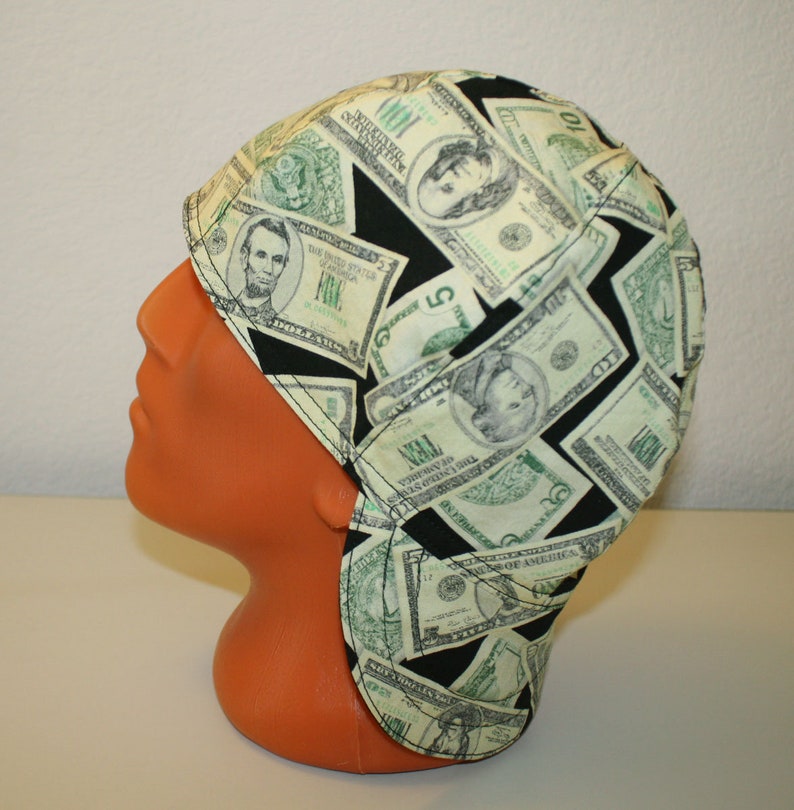 May include: A welding cap featuring a repeating pattern of US dollar bills in green and black. The cap is made of fabric and has a curved brim and ear flaps. The cap is displayed on an orange mannequin head.
