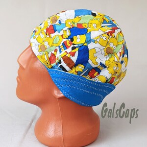 May include: A blue and white welding cap with a colorful cartoon print of the Simpsons characters. The cap has a blue brim and a white top with a zig-zag stitch around the edge. The text "GalsCaps" is visible in the lower right corner of the image.