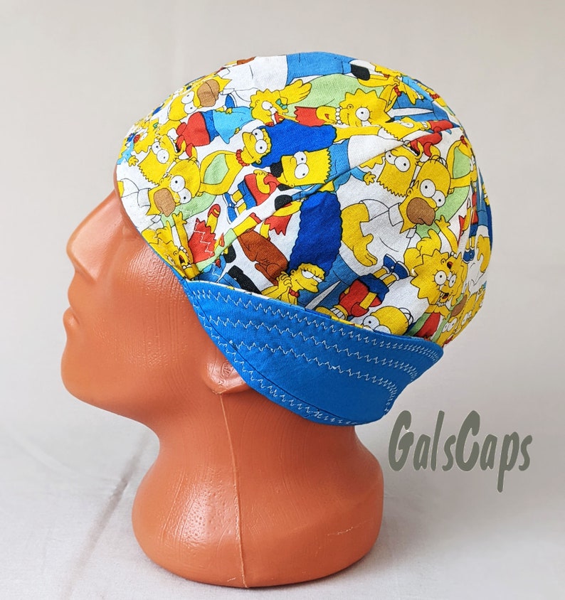 May include: A blue and white welding cap with a colorful cartoon print of the Simpsons characters. The cap has a blue brim and a white top with a zig-zag stitch around the edge. The text "GalsCaps" is visible in the lower right corner of the image.