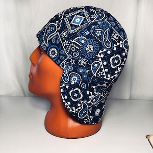 May include: A blue and white paisley print welding cap. The cap is made of fabric and has ear flaps. The paisley pattern includes floral and geometric designs in shades of blue and white.