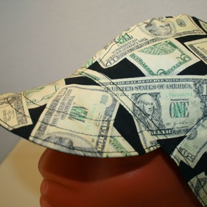 May include: A welding cap featuring a repeating pattern of US one-dollar bills and black geometric shapes. The cap is made of fabric and has a curved brim. The design includes the text "THE UNITED STATES OF AMERICA" and the number "ONE".