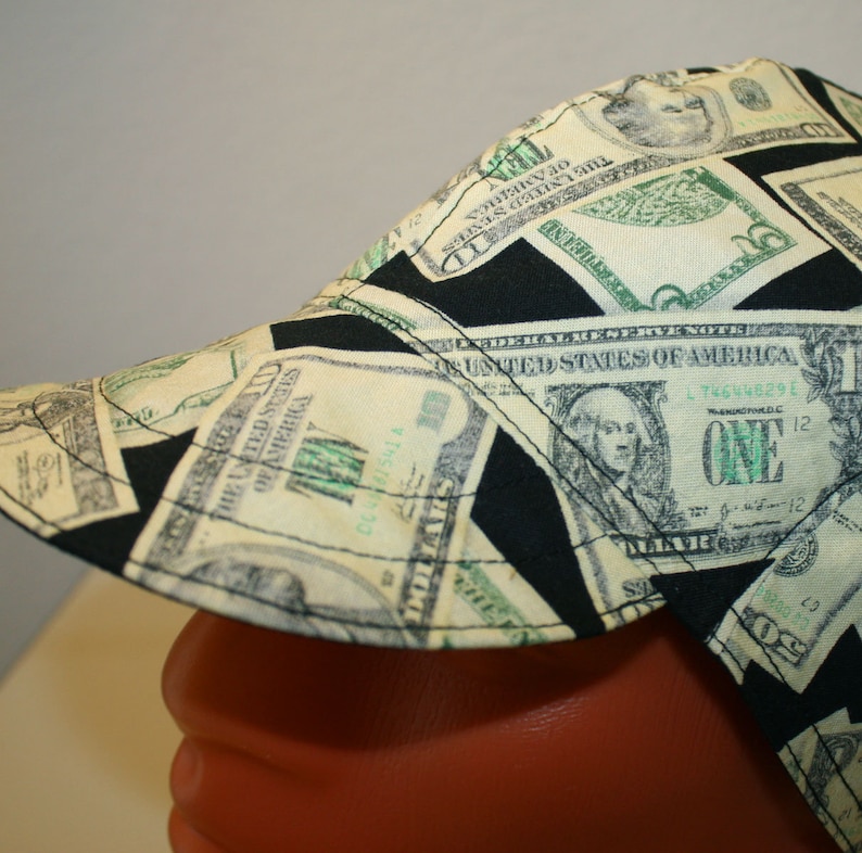 May include: A welding cap featuring a repeating pattern of US one-dollar bills and black geometric shapes. The cap is made of fabric and has a curved brim. The design includes the text "THE UNITED STATES OF AMERICA" and the number "ONE".