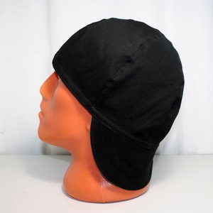May include: A black cap with ear flaps, displayed on a mannequin head. The cap is made of a solid, dark fabric and has a rounded crown. The ear flaps extend down to cover the ears. The cap is designed to provide warmth and protection.