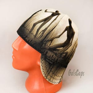 May include: A welding cap featuring a graphic print of two tall, slender, dark humanoid figures standing in a misty, muted forest setting. The cap has a black stitched brim and a small black fabric tag with the text GalsCaps on the side.