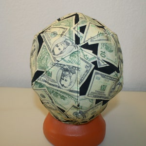 May include: A fabric cap featuring a repeating pattern of U.S. dollar bills in shades of green and black. The cap is resting on an orange form, showcasing the design.