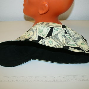 May include: A black and money-print cap. The cap features a black bill and a crown patterned with images of U.S. currency, including one, ten, and twenty dollar bills. The cap is displayed on a mannequin head.
