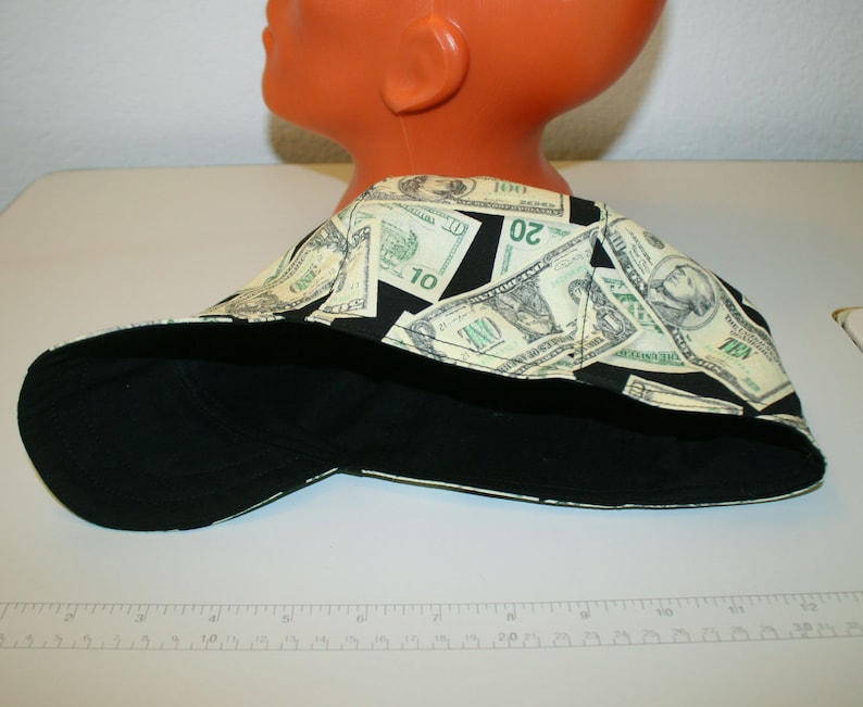 May include: A black and money-print cap. The cap features a black bill and a crown patterned with images of U.S. currency, including one, ten, and twenty dollar bills. The cap is displayed on a mannequin head.