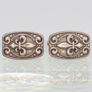 Puede incluir: Un par de gemelos plateados con un diseño ornamentado. Cada gemelo presenta un motivo central de flor de lis, rodeado de patrones arremolinados y pequeños detalles de cuentas dentro de un marco rectangular. Los gemelos se muestran sobre una superficie reflectante.
