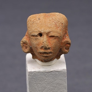 May include: A small, ancient-looking terracotta head sculpture. The head has a weathered appearance with visible details of the face, including closed eyes, a defined nose, and a mouth. The hair is styled with side curls. It is mounted on a white block.