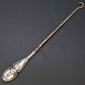 May include: A vintage silver crochet hook with a decorative handle featuring an embossed floral design. The hook has a slender, elongated shaft and a curved end. The hook is approximately 7 inches long.