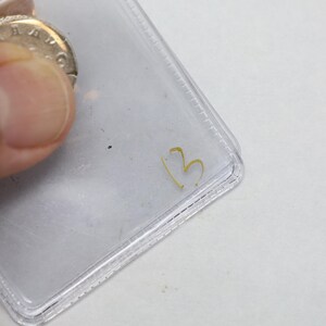 K&ouml;nnte beinhalten: Eine silberne M&uuml;nze ist teilweise sichtbar, gehalten in einer transparenten Plastikh&uuml;lle. Auf der H&uuml;lle ist die goldene Zahl "3" geschrieben. Die Details der M&uuml;nze sind teilweise verdeckt.