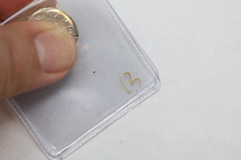 K&ouml;nnte beinhalten: Eine silberne M&uuml;nze ist teilweise sichtbar, gehalten in einer transparenten Plastikh&uuml;lle. Auf der H&uuml;lle ist die goldene Zahl "3" geschrieben. Die Details der M&uuml;nze sind teilweise verdeckt.