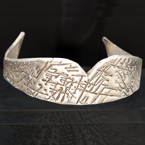 May include: Silver-toned cuff bracelet with a textured surface and geometric patterns. The bracelet has a wide band with raised edges and a central V-shaped dip. The design is abstract and modern.