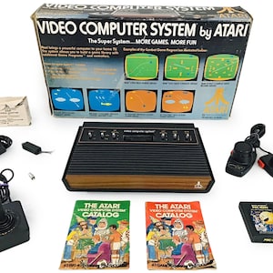 以下が含まれることがあります： Atari Video Computer Systemゲーム機セット。 セットには、コンソール、コントローラー、ゲームカートリッジ、カタログが含まれます。 コンソールは黒色で、木目調の仕上げです。 箱にはゲームのスクリーンショットとAtariのロゴが表示されています。
