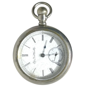 Può includere: Un orologio da tasca argentato con quadrante bianco e numeri romani neri. L'orologio ha un piccolo quadrante dei secondi e una lunetta strutturata. L'orologio è sospeso da un anello d'argento nella parte superiore. Il quadrante dell'orologio ha la scritta "Elgin Natl Watch Co."