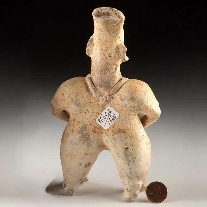 May include: A weathered, tan-coloured figurine with a tall, cylindrical head and arms behind its back. The figure has a rounded body and legs, with a small tag attached. A penny is placed nearby for scale, showing the size of the figurine.