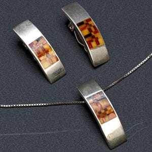 Puede incluir: Un conjunto de collar con colgante y pendientes a juego de plata. Los colgantes y pendientes rectangulares presentan un mosaico de tonos ámbar, naranja y marrón. Las joyas se exhiben sobre una superficie gris oscuro.