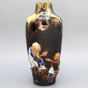 Könnte beinhalten: Eine hohe, dunkelbraune Keramikvase mit strukturierter Oberfläche und glänzender, getropfter Glasur in Beige- und Brauntönen. Die Vase zeigt eine Reliefskulptur von zwei Figuren in traditioneller Kleidung. Die Figuren sind in Weiß-, Blau- und Brauntönen gehalten.