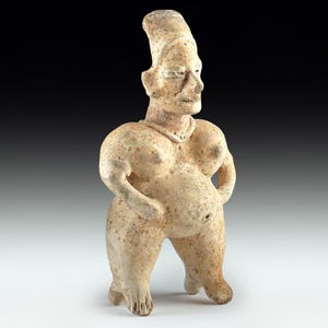 May include: A beige-coloured, ancient-style figurine of a standing figure with a large belly and prominent breasts. The figure has a tall, cylindrical headdress and arms at its sides. The sculpture is made of a textured material, possibly clay or stone.