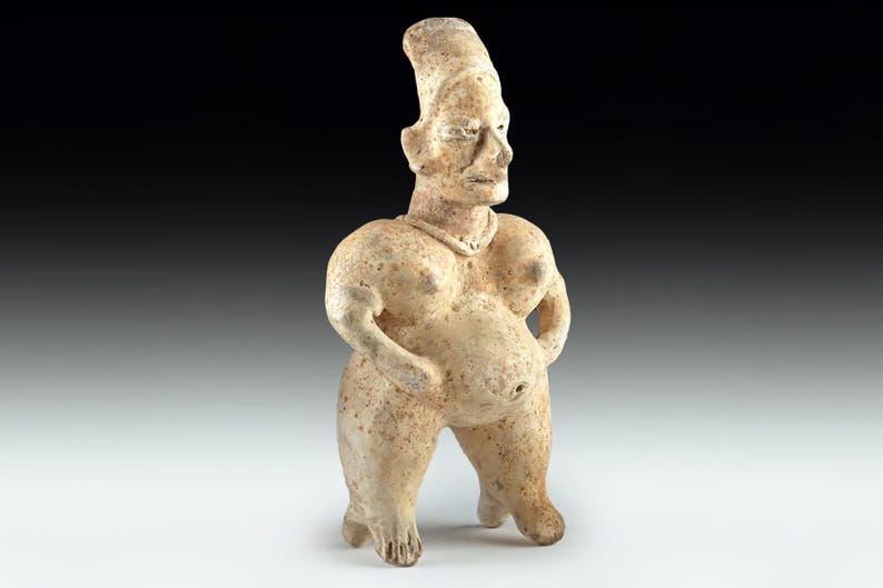 May include: A beige-coloured, ancient-style figurine of a standing figure with a large belly and prominent breasts. The figure has a tall, cylindrical headdress and arms at its sides. The sculpture is made of a textured material, possibly clay or stone.