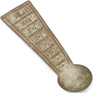 Op de afbeelding: Een langwerpig metalen object in bronskleur met een cirkelvormig uiteinde. Het hoofdgedeelte heeft rechthoekige panelen met geëtste halve maan- en stippatronen. Het object heeft een verweerde uitstraling.