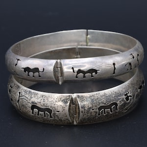 Op de afbeelding: Twee zilverkleurige scharnierende armbanden. Elke armband heeft uitgesneden ontwerpen van dieren en figuren. De armbanden worden gepresenteerd op een donkere ondergrond, die hun ingewikkelde details en vintage stijl benadrukt. Een uniek accessoire.