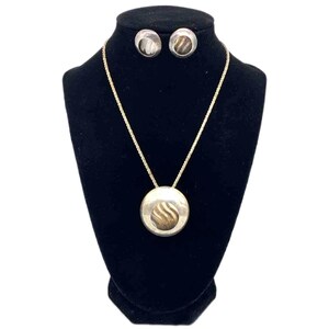 May include: A silver-toned jewelry set displayed on a black mannequin. The set includes a necklace with a circular pendant and matching stud earrings. The pendant and earrings feature a textured, swirling design in a contrasting color.