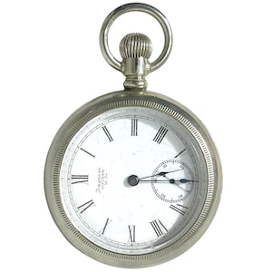 Può includere: Un orologio da tasca argentato con quadrante bianco e numeri romani neri. L'orologio ha un piccolo quadrante dei secondi e un anello nella parte superiore per una catena. Le parole "American Waltham W. Co." sono stampate sul quadrante.