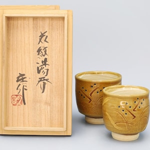 May include: Two small, tan ceramic teacups with a decorative pattern, presented with a wooden box. The box has Japanese calligraphy and a red stamp. The teacups have a light-coloured base.
