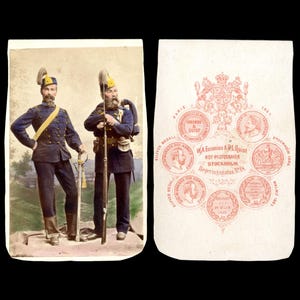 May include: A vintage photograph featuring two men in military uniforms, standing side-by-side. The men are wearing dark blue jackets, pants, and hats with yellow accents. One holds a sword, the other a rifle. The reverse side has a decorative design with text.