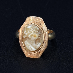 Puede incluir: Anillo dorado con un camafeo ovalado que representa un retrato de perfil. El camafeo está engastado en un marco decorativo texturizado. La banda del anillo es de un color dorado más oscuro.