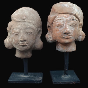 Puede incluir: Dos esculturas antiguas de terracota de cabezas humanas, cada una montada sobre un soporte de metal negro. Las cabezas tienen los ojos cerrados y rasgos faciales detallados. Una es de color beige claro y la otra marrón rojizo.