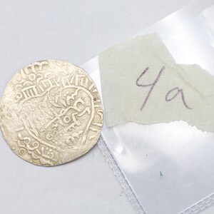 Puede incluir: Una moneda de plata con intrincados dise&ntilde;os y s&iacute;mbolos en relieve. La moneda es redonda y parece antigua. Un trozo de papel rasgado con el texto manuscrito "4a" est&aacute; junto a la moneda, dentro de una bolsa de pl&aacute;stico transparente.