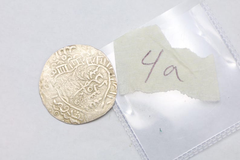 Puede incluir: Una moneda de plata con intrincados dise&ntilde;os y s&iacute;mbolos en relieve. La moneda es redonda y parece antigua. Un trozo de papel rasgado con el texto manuscrito "4a" est&aacute; junto a la moneda, dentro de una bolsa de pl&aacute;stico transparente.