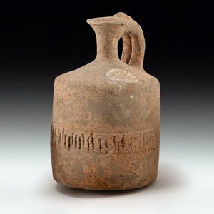 Może przedstawiać: Brązowy ceramiczny dzban w stylu antycznym, z uchwytem i wylewem. Dzban ma cylindryczny korpus z ozdobnym pasem pionowych linii. Wygląda na ręcznie robiony i ma rustykalny, postarzany wygląd.