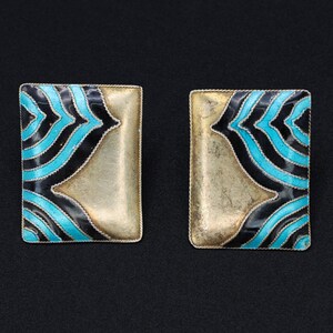 May include: Two rectangular earrings with a gold-colored base and a decorative pattern of black and turquoise blue curved lines. The earrings are set against a black background.