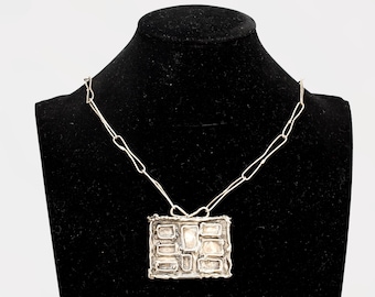 Puede incluir: Un collar plateado con un colgante rectangular. El colgante presenta un diseño abstracto con múltiples formas rectangulares y cuadradas. El collar se exhibe en un maniquí negro.