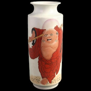Może przedstawiać: Biały ceramiczny wazon z malowaną ilustracją uśmiechniętej postaci Buddy w czerwonych szatach, trzymającej laskę. Tło przedstawia różową kulę i beżową podstawę.