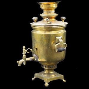 May include: A vintage brass samovar with a spigot, handle, and decorative top. The tea urn has a cylindrical body and sits on a square base. The samovar is a golden color and is set against a black background.