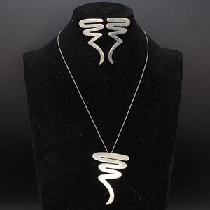 Puede incluir: Un conjunto de joyas plateadas que incluye un collar y pendientes a juego. El collar tiene una cadena larga y fina con un colgante abstracto estilizado. Los pendientes tienen un diseño similar. Las joyas se exhiben sobre un busto negro.