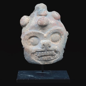 Puede incluir: Una escultura de piedra gris desgastada de una cara estilizada con una amplia sonrisa dentuda y ojos grandes y redondos. La escultura tiene protuberancias bulbosas en la parte superior y está montada sobre un soporte negro, sobre un fondo negro.
