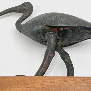 Peut inclure: Sculpture d'oiseau en bronze avec un long bec incurv&eacute; et des pattes fines. L'oiseau est pos&eacute; sur une base rectangulaire en bois. La sculpture a un aspect vieilli avec des reflets verts et rouges.
