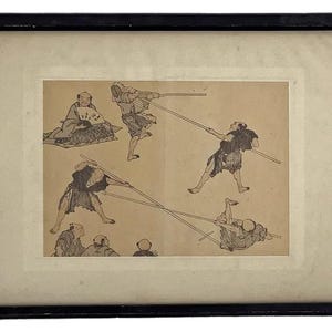 May include: Framed vintage print featuring several figures engaged in a martial arts practice with long poles. The artwork is in black and white, with a sepia-toned background. The print is set in a black frame with a white mat.