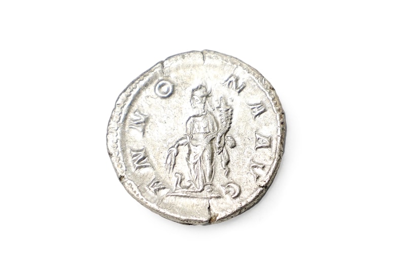 K&ouml;nnte beinhalten: Eine silberne r&ouml;mische M&uuml;nze mit einem detaillierten Relief einer Figur, die einen Stab und ein Weizenb&uuml;ndel h&auml;lt. Die M&uuml;nze hat sichtbare Inschriften am Rand und ist vor einem wei&szlig;en Hintergrund platziert. Die M&uuml;nze ist leicht abgenutzt.