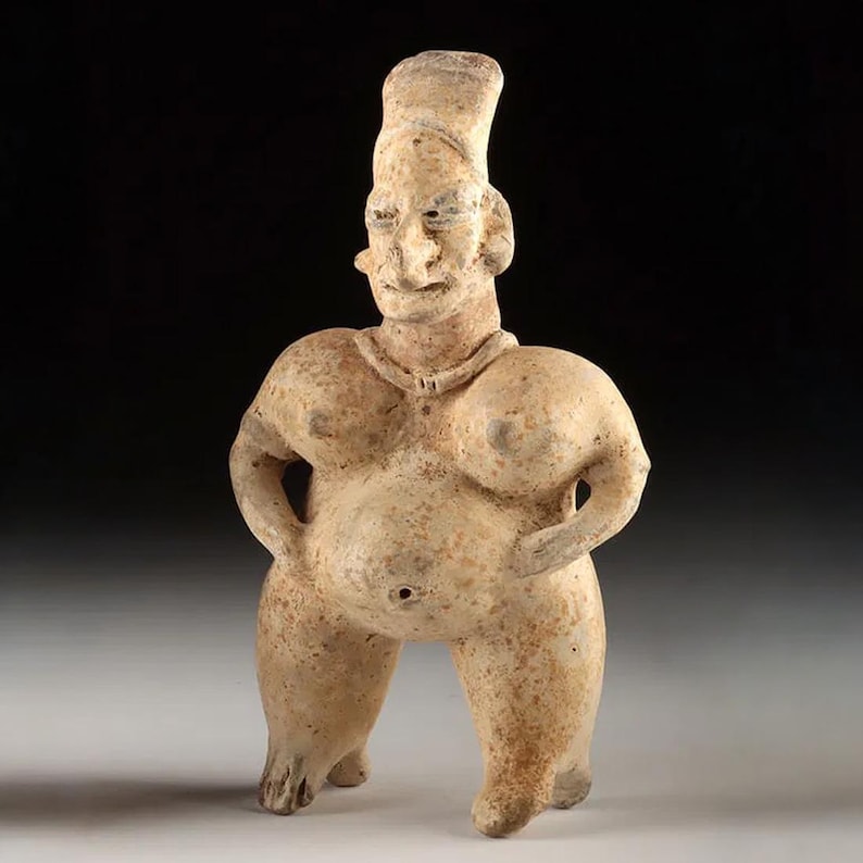 May include: A tan-coloured, ancient-style figurine of a person with a large belly and breasts. The figure has a tall, cylindrical headdress and is standing with hands on hips. The sculpture is made of a clay-like material and has a weathered appearance.