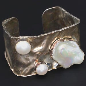 May include: A silver cuff bracelet with a hammered texture. The bracelet features three white pearls, including a large, irregularly shaped pearl. The jewelry is set against a dark background.