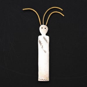 May include: A silver pendant in the shape of a stylized figure with two small eye holes and three gold-colored curved lines extending from the head. The figure has etched details on the front and is set against a black background.